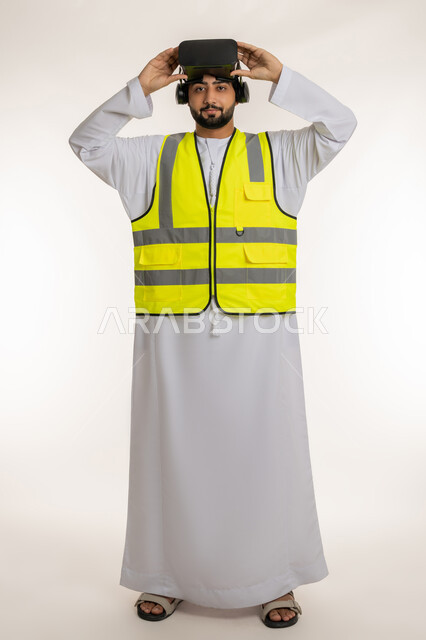 تطور المجال الهندسي، التفاعل مع الهندسة الافتراضية، بورتريه لمهندس عربي خليجي إماراتي يرتدي الكندورة والسترة الخاصة بالعمل يضع نظارة الواقع الافتراضي على رأسه ينظر إلى الكاميرا بإيماءات الثقة، دمج التكنولوجيا بالمجالات الهندسية، الجسم بالطول الكامل، خلفية بيضاء