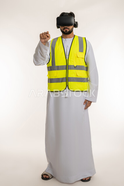 تجسيد ومشاهدة المشاريع الهندسية بالاعتماد على التكنولوجيا، الاندماج مع الهندسة الافتراضية، بورتريه لمهندس عربي خليجي إماراتي يرتدي الكندورة والسترة الخاصة بالعمل ونظارة الواقع الافتراضي يرفع إصبع السبابة للمس عناصر هولوجرامية، الجسم بالطول الكامل، خلفية بيضاء