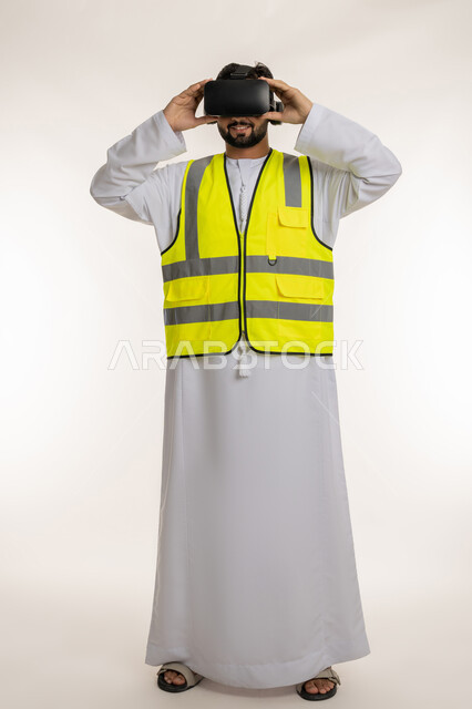 مشاهدة المشاريع الهندسية بالاعتماد على التكنولوجيا، التفاعل مع الهندسة الافتراضية، بورتريه لمهندس عربي خليجي إماراتي يرتدي الكندورة والسترة الخاصة بالعمل ونظارة الواقع الافتراضي، تطور المجال الهندسي في الإمارات العربية المتحدة، الجسم بالطول الكامل، خلفية بيضاء