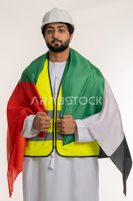 النظر إلى الكاميرا بإيماءات الاعتزاز بالانتماء للوطن، يوم العلم الإماراتي 3 نوفمبر، ذكرى قيام الاتحاد 2 ديسمبر 1971، بورتريه لمهندس عربي خليجي إماراتي يرتدي الكندورة والخوذة وسترة الحماية يضع على كتفه علم الإمارات المتحدة، العمل في القطاع الهندسي، خلفية بيضاء