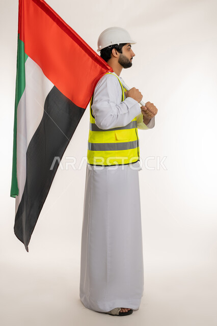 إيماءات الفخر والاعتزاز بالانتماء للوطن، ذكرى قيام الاتحاد 2 ديسمبر 1971، بورتريه من الجانب لمهندس عربي خليجي إماراتي يرتدي الكندورة والخوذة وسترة الحماية يحمل علم دولة الإمارات المتحدة، يوم العلم الإماراتي 3 نوفمبر، الجسم بالطول الكامل، خلفية بيضاء