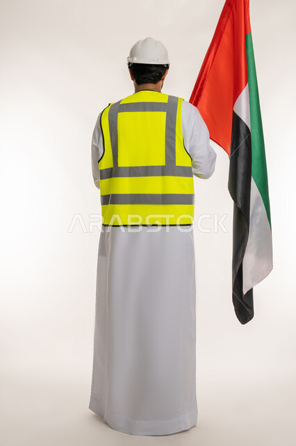 يوم العلم الإماراتي 3 نوفمبر، ذكرى قيام الاتحاد 2 ديسمبر 1971، بورتريه من الخلف لمهندس عربي خليجي إماراتي يرتدي الكندورة والخوذة وسترة الحماية يحمل علم دولة الإمارات المتحدة، العمل في القطاع الهندسي، الاعتزاز بالانتماء للوطن، الجسم بالطول الكامل، خلفية بيضاء