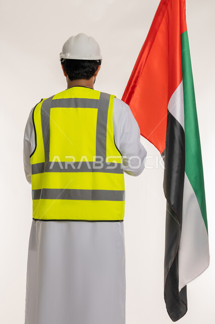 ذكرى قيام الاتحاد 2 ديسمبر 1971، بورتريه من الخلف لمهندس عربي خليجي إماراتي يرتدي الكندورة والخوذة وسترة الحماية يحمل علم دولة الإمارات المتحدة، العمل في القطاع الهندسي، الفخر والاعتزاز بالانتماء للوطن، يوم العلم الإماراتي 3 نوفمبر، خلفية بيضاء