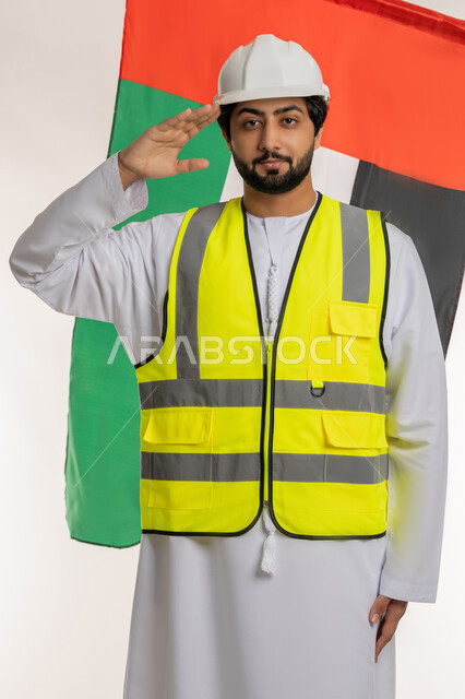 إلقاء التحية للوطن، النظر إلى الكاميرا بإيماءات الفخر والاعتزاز، يوم العلم الإماراتي 3 نوفمبر، ذكرى قيام الاتحاد 2 ديسمبر 1971، بورتريه لمهندس عربي خليجي إماراتي يرتدي الكندورة والخوذة وسترة الحماية خلفه علم الإمارات المتحدة، خلفية بيضاء