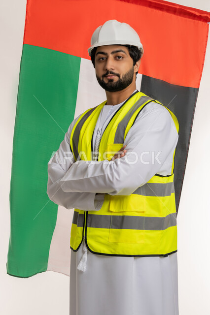 يوم العلم الإماراتي 3 نوفمبر، ذكرى قيام الاتحاد 2 ديسمبر 1971، بورتريه لمهندس عربي خليجي إماراتي يرتدي الكندورة والخوذة وسترة الحماية يقف بأيدي متقاطعة ينظر إلى الكاميرا بإيماءات الفخر خلفه علم دولة الإمارات العربية المتحدة، حب الوطن والانتماء إليه، خلفية بيضاء