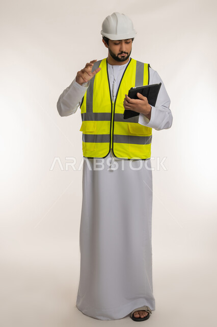 الأجهزة التقنية الحديثة، التأكد من سير الأعمال بالشكل المطلوب والصحيح، بورتريه لمهندس عربي خليجي إماراتي يرتدي الكندورة والخوذة وسترة الحماية يقوم باستخدام الجهاز اللوحي، تدوين و تسجيل الملاحظات, كتابة آخر المستجدات حول المشروع، الجسم بالطول الكامل، خلفية بيضاء