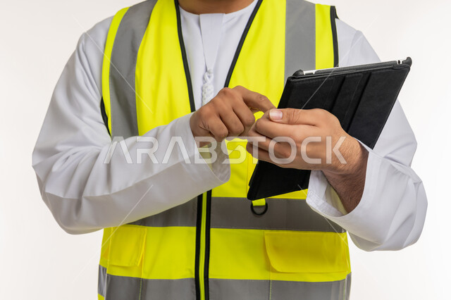 إدارة المشاريع الهندسية، التأكد من سير الأعمال بالشكل المطلوب، بورتريه مقرب لمهندس عربي خليجي إماراتي يرتدي الكندورة وسترة الحماية يقوم باستخدام الجهاز اللوحي، إنجاز مهام العمل عن بعد، الأجهزة التقنية الحديثة، تدوين وتسجيل الملاحظات, خلفية بيضاء