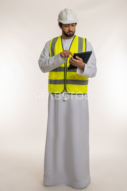 Taking notes, writing down the latest updates on the project, ensuring that the work is proceeding as required and correctly, portrait of an Arab Gulf Emirati engineer wearing a kandura, helmet and protective jacket using a tablet, modern technical devices, full-length body, white background