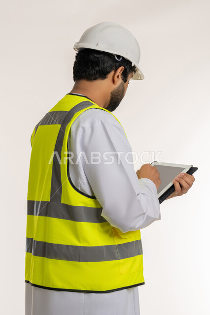 كتابة آخر المستجدات حول المشروع، الأجهزة التقنية الحديثة، التأكد من سير الأعمال بالشكل المطلوب والصحيح، بورتريه من الخلف لمهندس عربي خليجي إماراتي يرتدي الكندورة وسترة الحماية يقوم باستخدام الجهاز اللوحي، تدوين و تسجيل الملاحظات, خلفية بيضاء