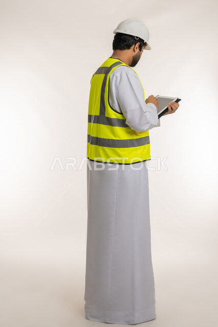 Completing work tasks remotely, modern technical devices, ensuring that work is proceeding as required, portrait from the back of an Arab Gulf Emirati engineer wearing a kandura, helmet and protective vest using a tablet, taking notes, full body, white background