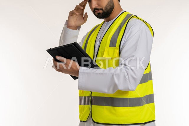 إدارة المشاريع الهندسية، بورتريه مقرب لمهندس عربي خليجي إماراتي يرتدي الكندورة وسترة الحماية يقوم باستخدام الجهاز اللوحي يضع يده على رأسه بإيماءات التفكير، إنجاز مهام العمل عن بعد، محاولة تذكر شيء ما، أجهزة تقنية حديثة، تدوين وتسجيل الملاحظات, خلفية بيضاء