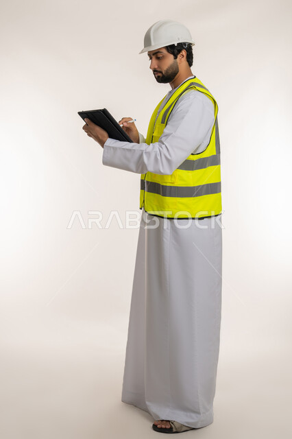 التأكد من سير الأعمال بالشكل المطلوب، بورتريه من الجانب لمهندس عربي خليجي إماراتي يرتدي الكندورة والخوذة وسترة الحماية يقوم باستخدام الجهاز اللوحي، إنجاز مهام العمل عن بعد، الأجهزة التقنية الحديثة، تدوين و تسجيل الملاحظات, الجسم بالطول الكامل، خلفية بيضاء