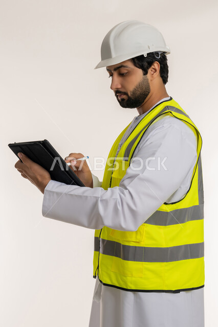 Engineering project management, ensuring work is going as required, side portrait of an Arab Gulf Emirati engineer wearing a kandura, helmet and safety vest using a tablet, completing work tasks remotely, modern technical devices, taking and recording notes, white background