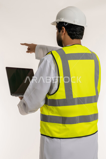 استخدام الأجهزة التقنية الحديثة، بورتريه من الخلف لمهندس عربي خليجي امارتي يرتدي الكندورة والخوذة وسترة الحماية يستخدم الحاسوب المحمول يشير بإصبع السبابة إلى شيء ما، إدارة المشاريع المعمارية والهندسية، إنجاز مهام العمل عن بعد، مهن ووظائف شبابية، خلفية بيضاء