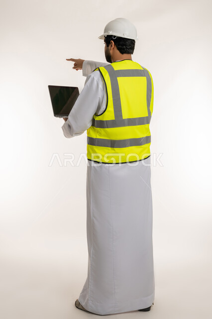 إنجاز مهام العمل عن بعد، الأجهزة التقنية الحديثة، بورتريه من الخلف لمهندس عربي خليجي امارتي يرتدي الكندورة والخوذة وسترة الحماية يستخدم الحاسوب المحمول يشير بإصبع السبابة إلى شيء ما، إدارة المشاريع الهندسية، مهن ووظائف شبابية، الجسم بالطول الكامل، خلفية بيضاء