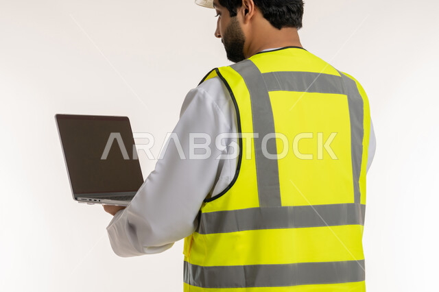 Architectural and engineering project management, using modern technical devices, close-up portrait from back of an Arab Gulf Emirati engineer wearing a kandura and a protective vest using a laptop, completing work tasks remotely, youth professions and jobs in the United Arab Emirates, white background