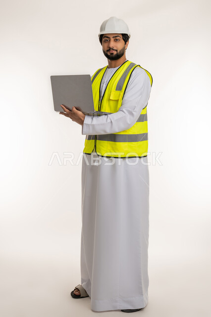 إنجاز مهام العمل عن بعد، استخدام الأجهزة التقنية الحديثة، بورتريه لمهندس عربي خليجي امارتي يرتدي الكندورة والخوذة وسترة الحماية يستخدم الحاسوب المحمول ينظر إلى الكاميرا، متابعة المشاريع المعمارية والهندسية، مهن ووظائف شبابية، الجسم بالطول الكامل، خلفية بيضاء