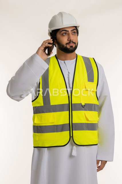 العمل في القطاع الهندسي في الإمارات العربية المتحدة, بورتريه لمهندس عربي خليجي امارتي يرتدي الكندورة والخوذة وسترة الحماية يحمل الهاتف المحمول يقوم بإجراء مكالمة هاتفية، استخدام الأجهزة التقنية الحديثة، متابعة العمل عن بعد، المهن والوظائف الشبابية، خلفية بيضاء