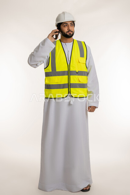 Youth jobs and professions, working in the engineering sector, portrait of an Arab Gulf Emirati engineer wearing a kandura, helmet and protective vest, holding a mobile phone making a phone call, using modern technical devices, following up on work remotely, full body length, white background