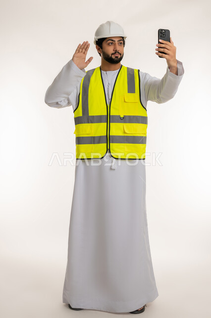 Raising hand to greet and welcome, working in the engineering sector, portrait of an Arab Gulf Emirati engineer wearing a kandura, helmet and protective vest holding a mobile phone making a video call, using modern technical devices, youth professions and jobs, full body, white background