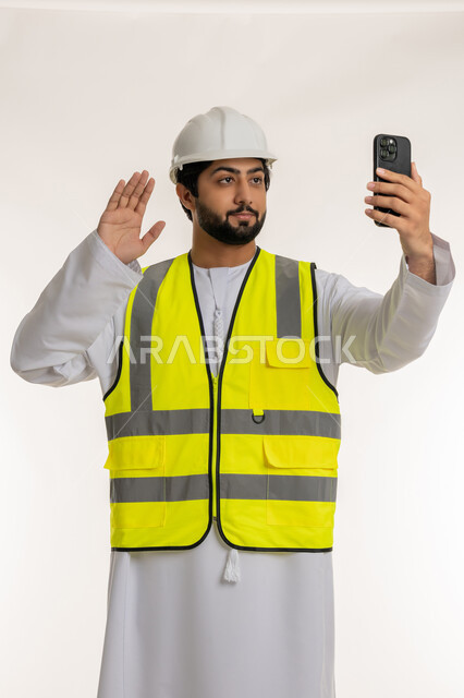 استخدام الأجهزة التقنية الحديثة، رفع اليد لإلقاء التحية والترحيب، العمل في القطاع الهندسي, بورتريه لمهندس عربي خليجي امارتي يرتدي الكندورة والخوذة وسترة الحماية يحمل الهاتف المحمول يقوم بإجراء مكالمة فيديو، المهن والوظائف الشبابية في الإمارات، خلفية بيضاء
