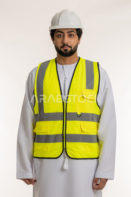 Concept of architectural construction and engineering, portrait of an Arab Gulf Emirati engineer wearing a kandura, helmet and protective vest standing straight looking at the camera with gestures of seriousness and confidence, youth professions and jobs in the United Arab Emirates, white background