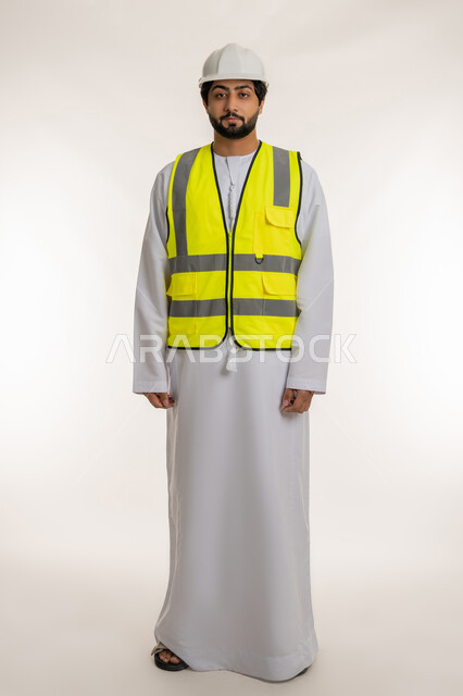 Youth professions and jobs in the Emirates, concept of architectural construction and engineering, portrait of an Arab Gulf Emirati engineer wearing a kandura, helmet and protective vest standing straight looking at the camera with gestures of seriousness and confidence, full body, white background