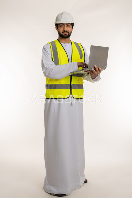Completing work tasks remotely, using modern technical devices, portrait of an Arab Gulf Emirati engineer wearing a kandura, helmet and protective vest using a laptop looking at the camera, following up on architectural and engineering projects, youth professions and jobs, full body, white background