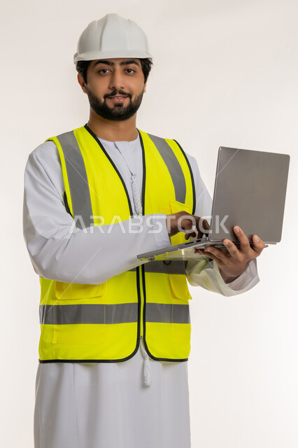 Follow up on architectural and engineering projects, use of modern technical devices, portrait of an Arab Gulf Emirati engineer wearing a kandura, helmet and protective vest using a laptop looking at the camera, completing work tasks remotely, youth professions and jobs in the Emirates, white background