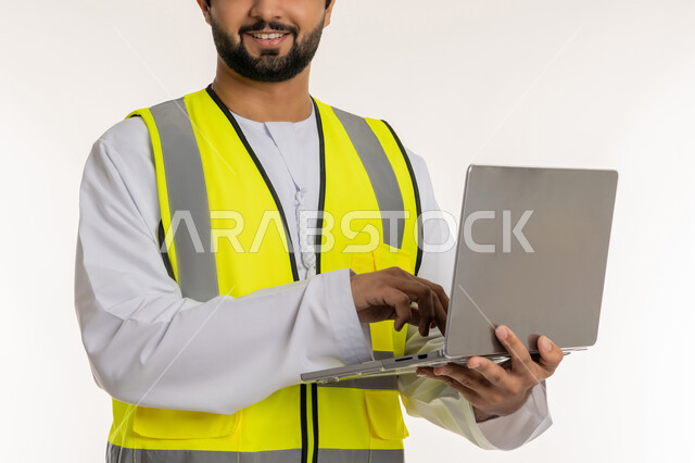 Completing work tasks remotely, managing architectural and engineering projects, using modern technical devices, close-up portrait of a smiling Emirati Gulf Arab engineer wearing a kandura and a protective vest using a laptop, youth professions and jobs in the United Arab Emirates, white background