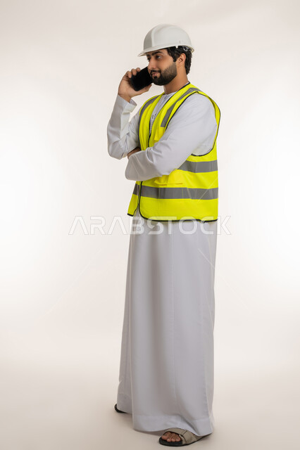 استخدام الأجهزة التقنية الحديثة، مهن ووظائف شبابية، العمل في القطاع الهندسي, بورتريه من الجانب لمهندس عربي خليجي امارتي يرتدي الكندورة والخوذة وسترة الحماية يحمل الهاتف المحمول يقوم بإجراء مكالمة هاتفية، متابعة العمل عن بعد، الجسم بالطول الكامل، خلفية بيضاء