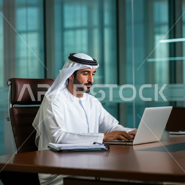 Using laptop to complete tasks, working in the administrative and office sector, professions and jobs for young people in the United Arab Emirates, a young Arab Gulf Emirati man wearing a kandura and a ghutra sitting behind his desk working hard and dedicatedly with gestures of integration and focus