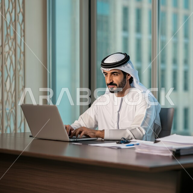 Using laptop to complete tasks, working in the administrative and office sector, professions and jobs for young people in the United Arab Emirates, a young Arab Gulf Emirati man wearing a kandura and a ghutra sitting behind his desk working hard and dedicatedly with gestures of integration and focus