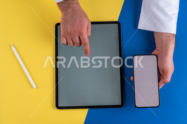 صورة مقربة ليد رجل عربي خليجي سعودي يحمل بيده الهاتف المحمول ، ينقر على شاشة جهاز التابلت، التسويق الالكتروني  ، تصفح المواقع الالكترونية ،اعلان ترويجي لتطبيق وموقع ويب ، استخدم التقنيات الحديثة في الاعمال الادارية