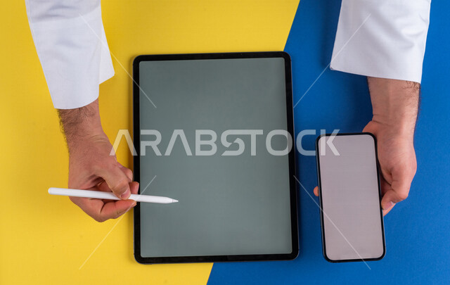صورة مقربة ليد رجل عربي خليجي سعودي يحمل الهاتف المحمول ويستخدام جهاز التابلت ، استخدام قلم التابلت لتدوين ملاحظات خاصة بالعمل ،شرح خطوات العمل ، اعلان ترويجي لتطبيق وموقع ويب ،استخدام التقنيات الحديثة في الاعمال الادارية