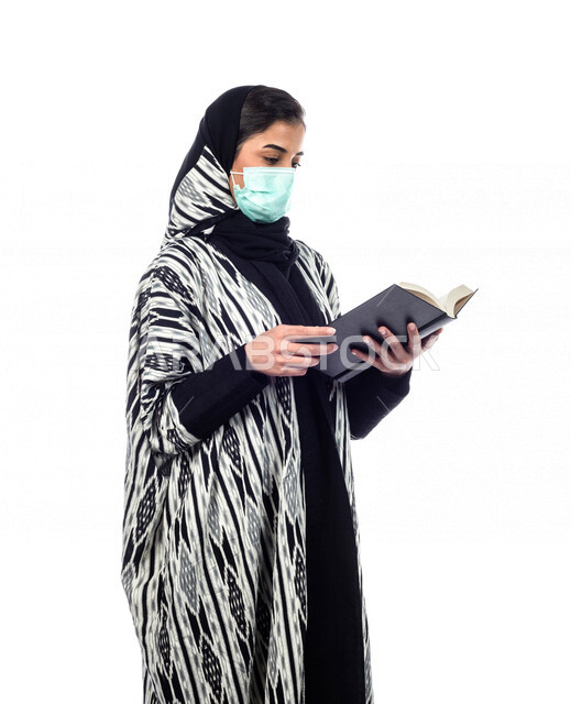 بورتريه لسيدة عربية خليجية ترتدي الكمامة الواقية، تقف وتحمل بيدها كتاب، قراءة الكتب، جلسة تصوير، الالتزام باجراءات الوقاية من فايروس كورونا، خلفية بيضاء