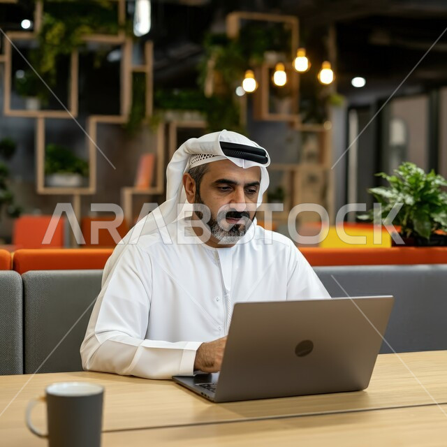Managing and organizing business affairs through modern technical devices, using advanced technology and techniques, completing work tasks remotely, a close-up image of an Emirati Gulf Arab man wearing a white kandura and ghutra sitting on the couch in an upscale cafe and using a laptop