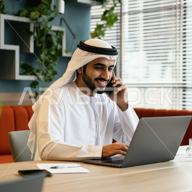 إدارة شؤون الأعمال وتنظيمها عبر الأجهزة التقنية الحديثة، استخدام التكنولوجيا والتقنيات المتطورة، إنجاز مهام العمل عن بعد، رجل عربي خليجي اماراتي يرتدي الكندورة والغترة البيضاء يجلس على الأريكة في إحدى المقاهي الراقية ويستخدم الحاسوب المحمول