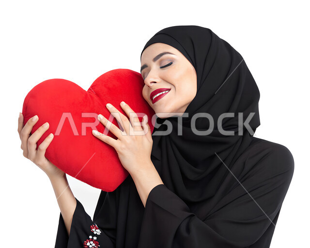 اظهار مشاعر الحب والشغف، مفهوم المحبة والمودة، بورتريه مقرب لامرأه عربيه خليجيه سعوديه محجبة ترتدي عباءة سوداء تحمل بيدها قلب باللون الاحمر بتعابير السرور والسعادة، خلفية بيضاء