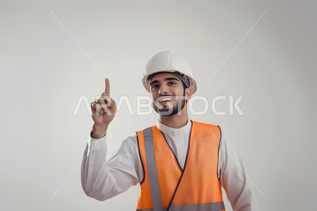 الإشارة بإصبع السبابة للأعلى, الإعمار بسواعد أبناء الوطن, مفهوم الهندسة والإنشاءات المعمارية, العمل في القطاع الهندسي, بورتريه مقرب لمهندس عربي خليجي سعودي يرتدي خوذة وسترة الحماية ينظر إلى الكاميرا بإيماءات السعادة والسرور, خلفية رمادية