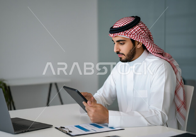 Integrating modern technology and technology into practical life, performing office tasks and work inside the company headquarters, close-up image of a Saudi Arabian Gulf man wearing traditional thobe and shemagh sitting in front of his desk holding a tablet, working in offices and companies in the Kingdom of Saudi Arabia