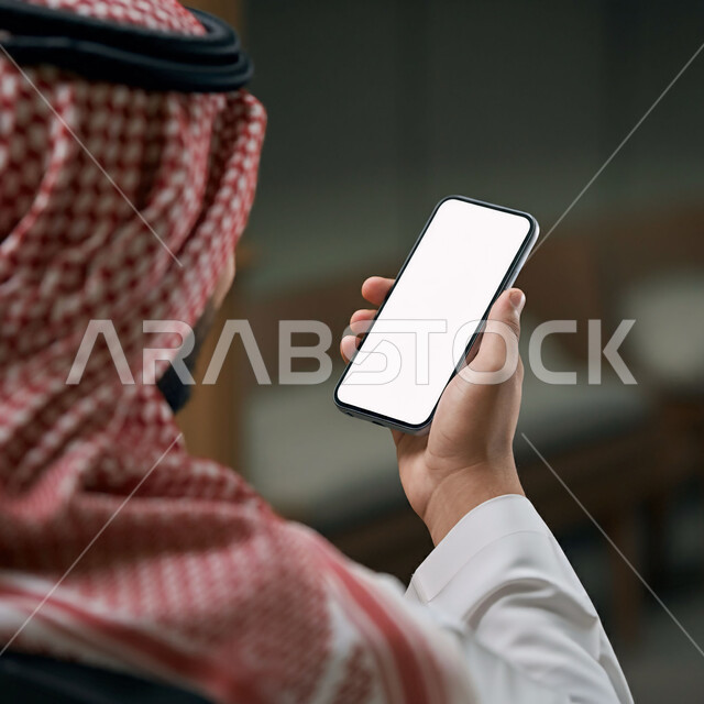 إدارة وإنجاز المهام عبر الأونلاين, صورة مقربة من الخلف لرجل عربي خليجي سعودي يرتدي الثوب التقليدي والشماغ يحمل بيده جوال بشاشة بيضاء فارغة, التسويق الرقمي والإعلانات التسويقية، استخدام الأجهزة والتقنيات الحديثة, التواصل مع الأهل عبر تطبيقات الهاتف الذكي