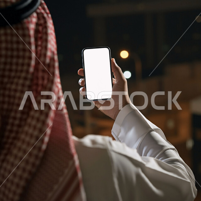 التسويق الرقمي والإعلانات التسويقية، استخدام الأجهزة والتقنيات الحديثة, إدارة وإنجاز المهام عبر الأونلاين, صورة مقربة من الخلف لرجل عربي خليجي سعودي يرتدي الثوب التقليدي والشماغ يحمل بيده جوال بشاشة بيضاء فارغة, التواصل مع الأهل عبر تطبيقات الهاتف الذكي
