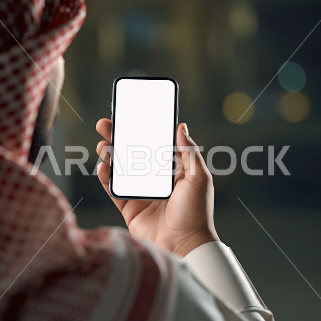 Using modern devices and technologies, managing and completing tasks online, a close-up image from the back of a Saudi Arabian Gulf man wearing a traditional thobe and shemagh holding a mobile phone with a blank white screen, communicating with family via smartphone applications, digital marketing and marketing advertisements