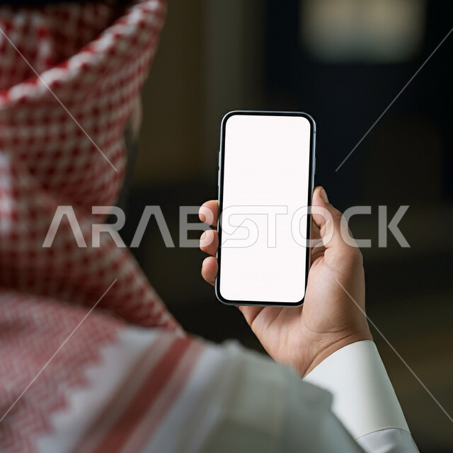 إدارة وإنجاز المهام عبر الأونلاين, التواصل مع الأهل عبر تطبيقات الهاتف الذكي, استخدام الأجهزة والتقنيات الحديثة, صورة مقربة من الخلف لرجل عربي خليجي سعودي يرتدي الثوب التقليدي والشماغ يحمل بيده جوال بشاشة بيضاء فارغة, التسويق الرقمي والإعلانات التسويقية