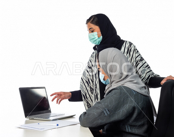 بورتريه لسيدة اعمال عربية خليجية سعودية تجلس امام الحاسوب المحمول، بجانبها موظفة خليجية سعودية، يقومان بمناقشة وتنظيم الاعمال، فريق عمل خليجي سعودي، ارتداء الكمامة للوقاية من فايروس كورونا