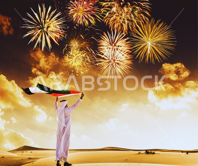 المفرقعات والألعاب النارية, الحب والانتماء للوطن, الاحتفال بالأعياد والمناسبات الوطنية, اليوم الوطني الإماراتي 1971, يوم العلم 3 نوفمبر, صورة من الخلف لرجل عربي خليجي إماراتي يرتدي الكندورة والغترة البيضاء يقف في الصحراء ويرفع علم الإمارات العربية المتحدة