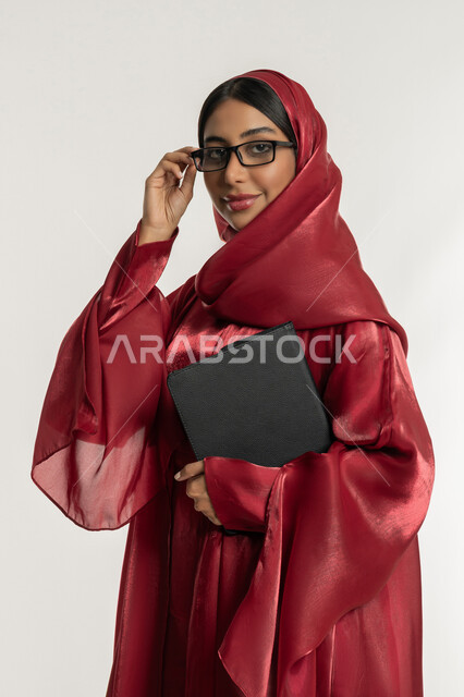 دمج التقنيات الحديثة والتكنولوجيا في الحياة العملية, بورتريه لامرأة عربية خليجية إماراتية محجبة ترتدي العباءة الحمراء تضع نظارة طبية تمسك بيدها الجهاز اللوحي, شابة سعودية تنظر إلى الكاميرا بإيماءات السعادة والسرور, خلفية بيضاء