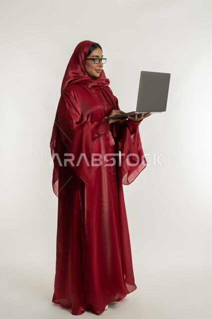 مفهوم العمل عن بعد, استخدام الأجهزة التقنية الحديثة والمتطورة, بورتريه لامرأة عربية خليجية إماراتية محجبة ترتدي العباءة الحمراء تضع نظارة طبية تعمل على جهاز الحاسوب المحمول, شابة سعودية تقوم بإنجاز المهام, الجسم بالطول الكامل, خلفية بيضاء