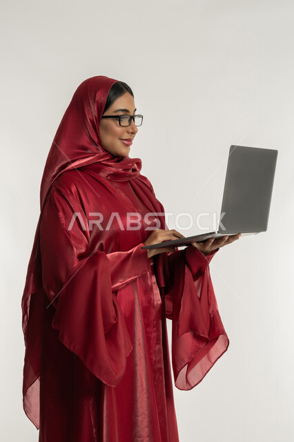 Using modern and advanced technical devices, the concept of remote work, a portrait of a veiled Emirati Gulf Arab woman wearing a red abaya, wearing medical glasses, working on a laptop, a young Saudi woman completing tasks, white background